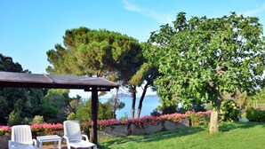 A patio with white chairs and a table, overlooking a body of water with trees and flowers in the background.