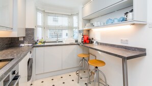 Een moderne keuken met witte kasten, een marmeren aanrechtblad en een inbouwoven. Op het aanrecht staan twee barkrukken en een koffieapparaat.