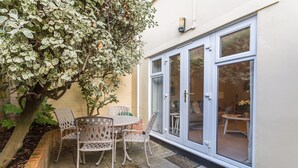 Een terras met een tafel, stoelen, een boom en een schuifdeur naar een binnenruimte.