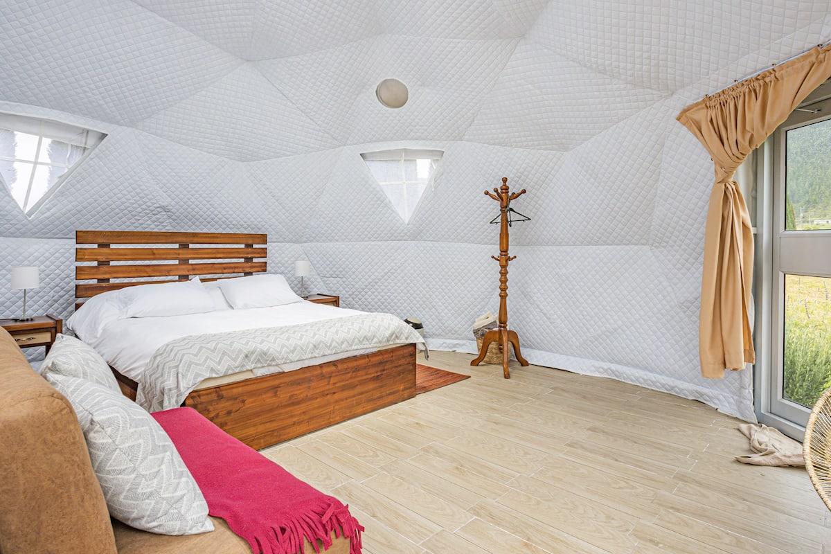 A bedroom with a wooden bed, a bedside table, a lamp, a coat rack, and a large window with curtains.