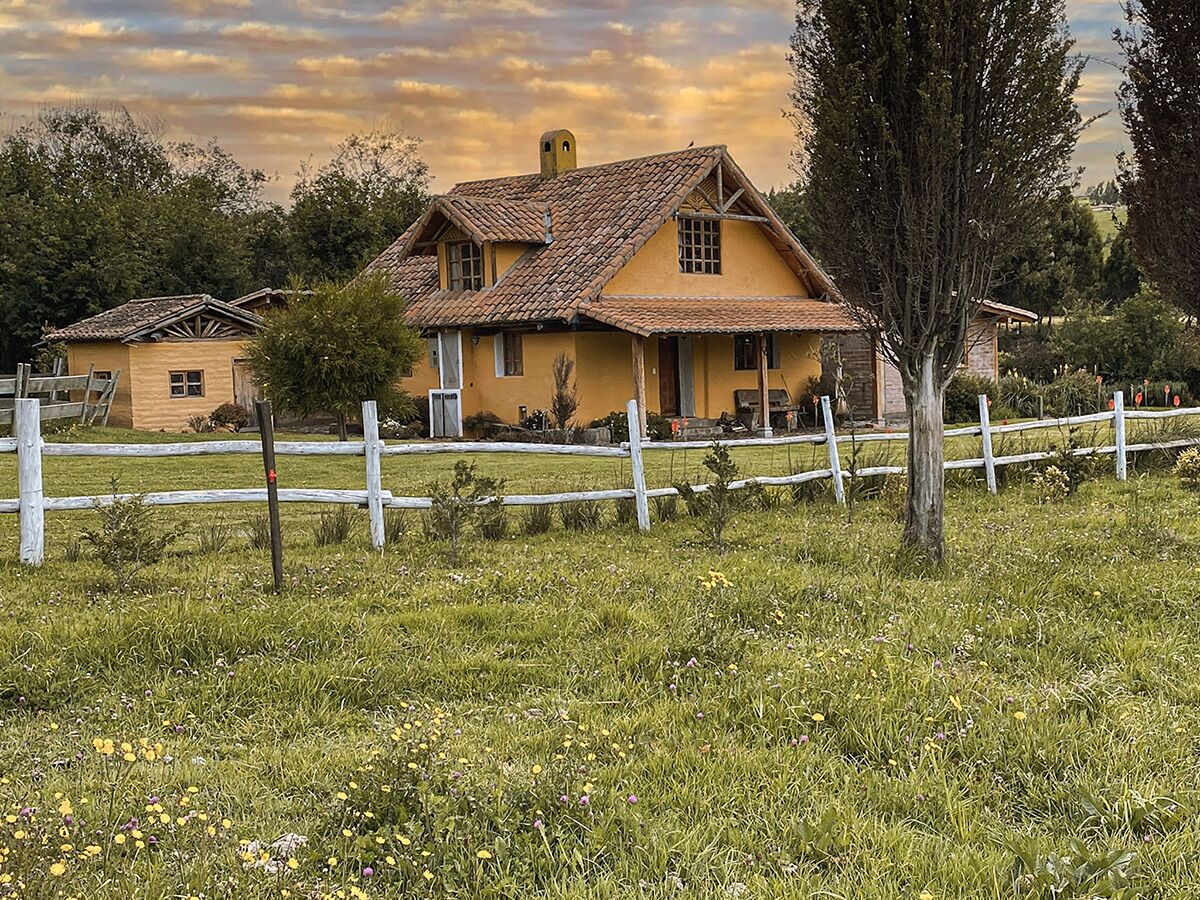 Una casa amarilla con techo de tejas, rodeada por una valla blanca y un campo cubierto de césped y flores silvestres.