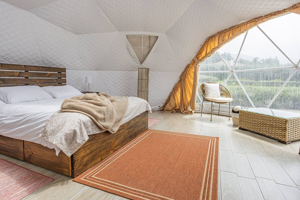A bedroom with a large bed, a wooden headboard, a bedside table with a lamp, a window with curtains, and a view of greenery outside.