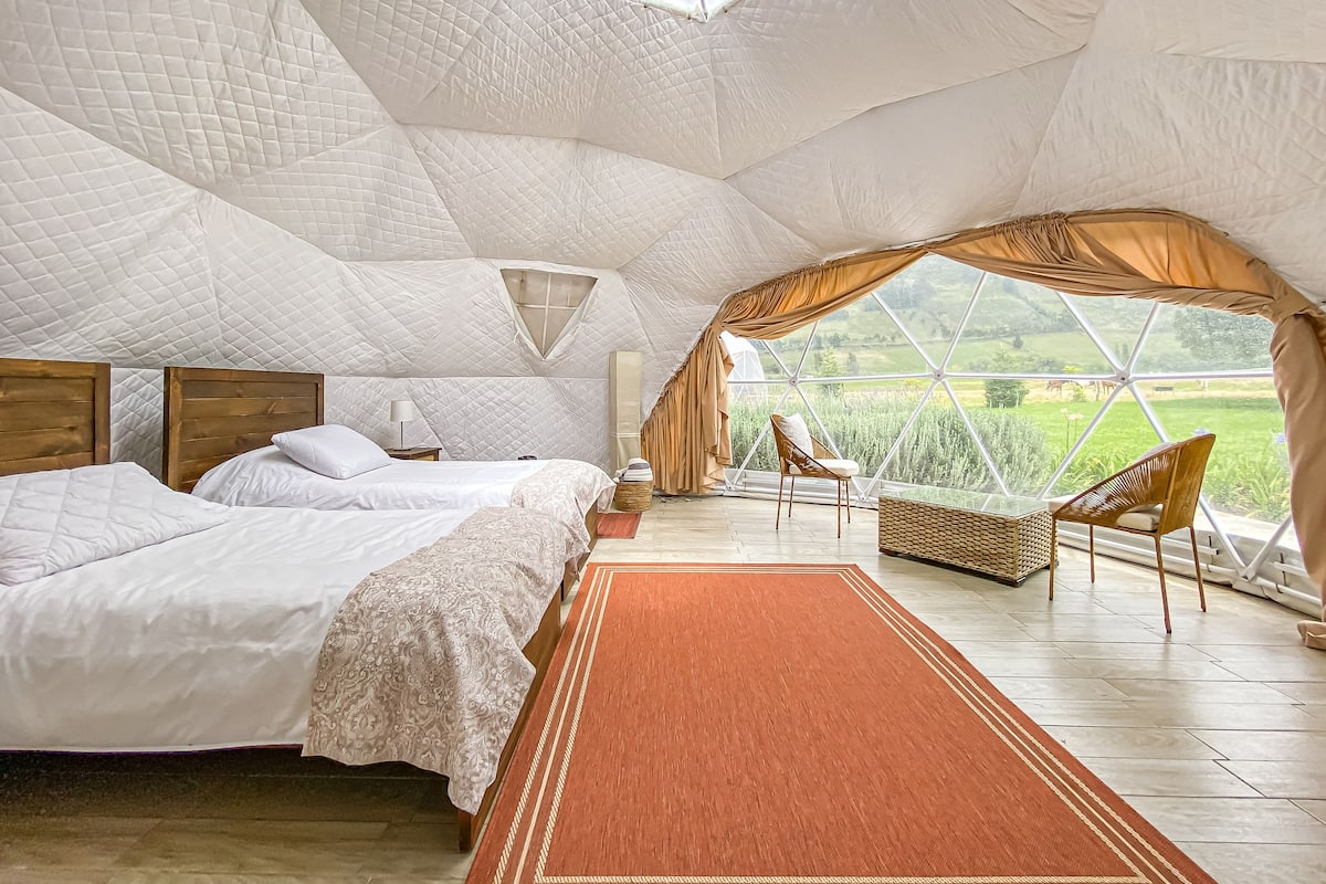 A spacious tented room with a large bed, a chair, and a view of a green field.