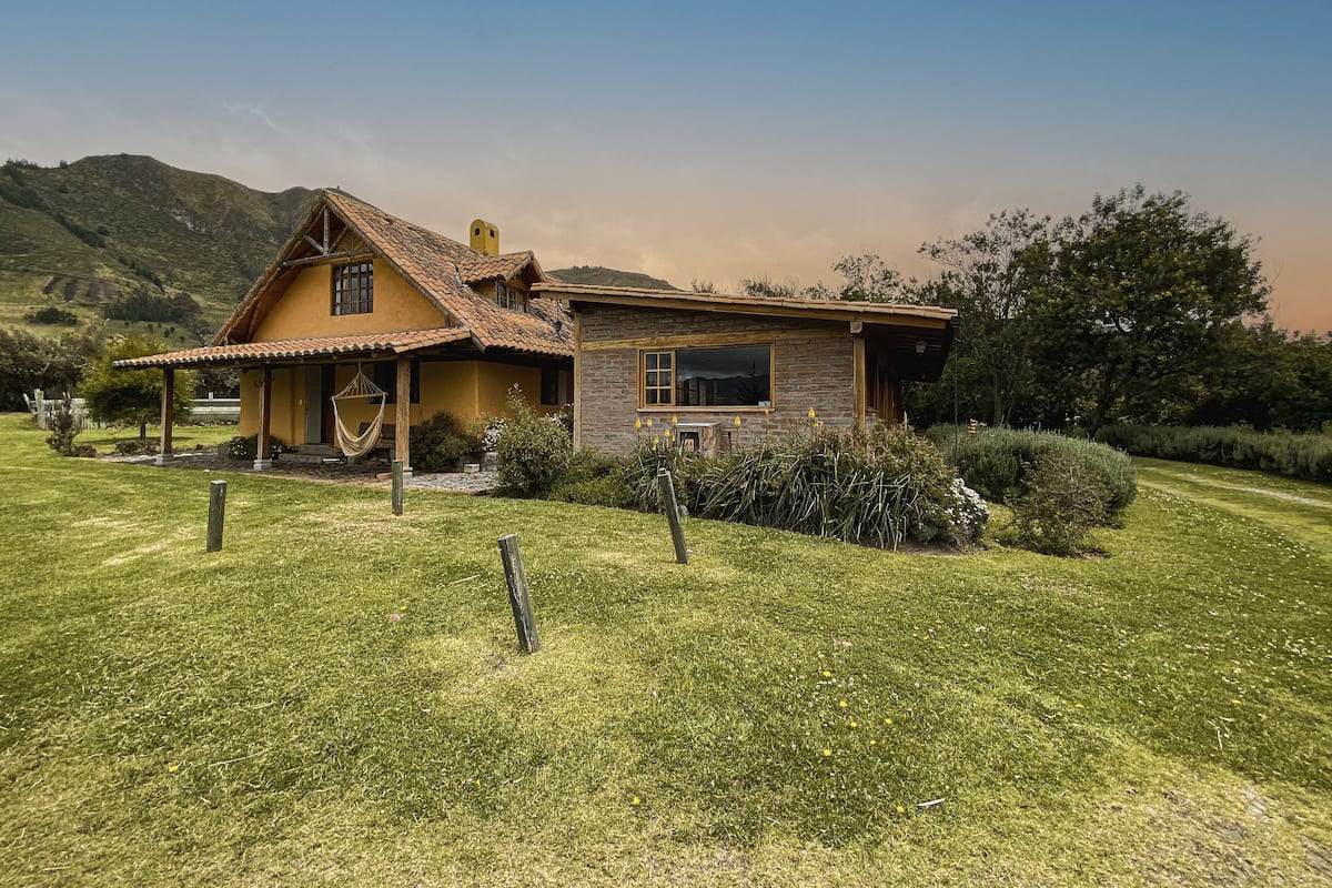 A rustic house with a stone facade and a tiled roof, surrounded by a well-maintained lawn and a mountain in the background.