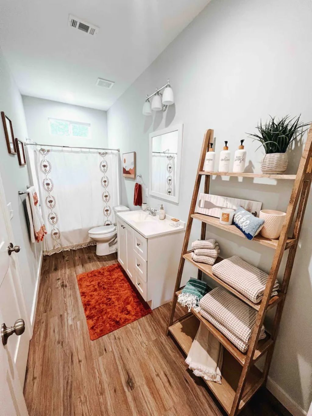 A modern bathroom with a wooden floor, white fixtures, a red rug, and a wooden shelf with towels and toiletries.