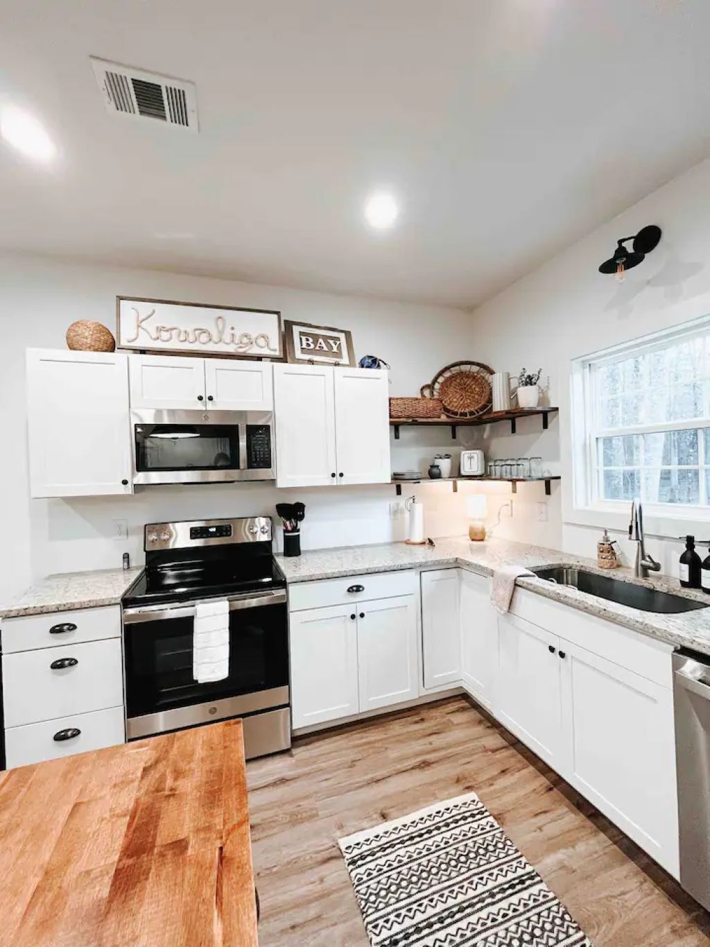 A modern kitchen with white cabinetry, a black stove, a microwave, and a sink. There are decorative items on the shelves and a window providing natural light.