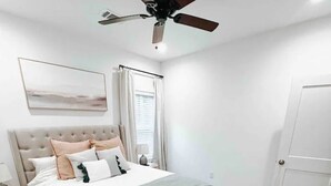 Quarto com cama, ventoinha no teto, mesa de cabeceira, abajur, janela com cortinas e porta.
