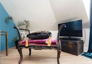 Un banc en bois foncé, doté d’un coussin moelleux et de quelques glands décoratifs, est placé dans une pièce dont le plafond est en pente et où se trouve un téléviseur posé sur un meuble.