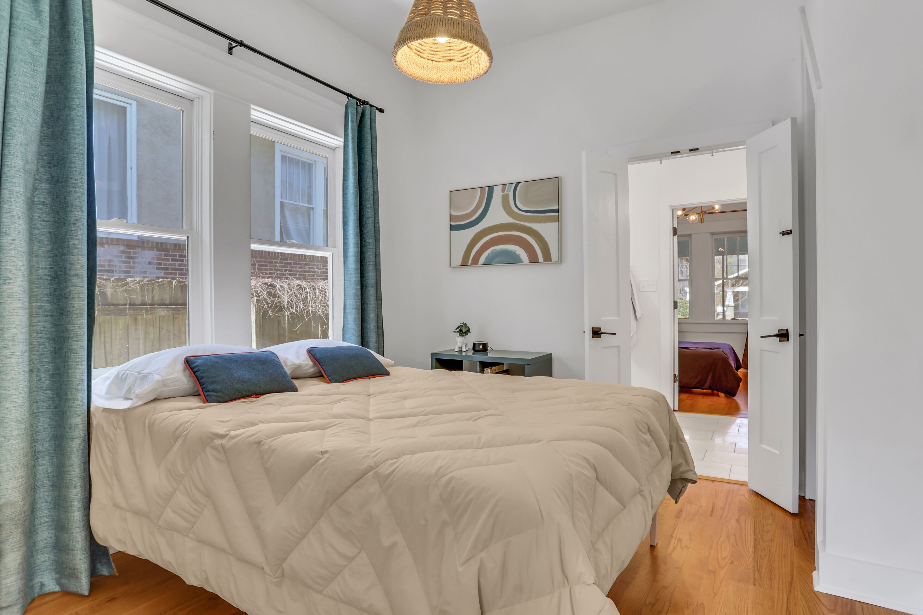 Une chambre avec un grand lit, une table de chevet, un luminaire suspendu et une vue sur une autre pièce par une porte ouverte.