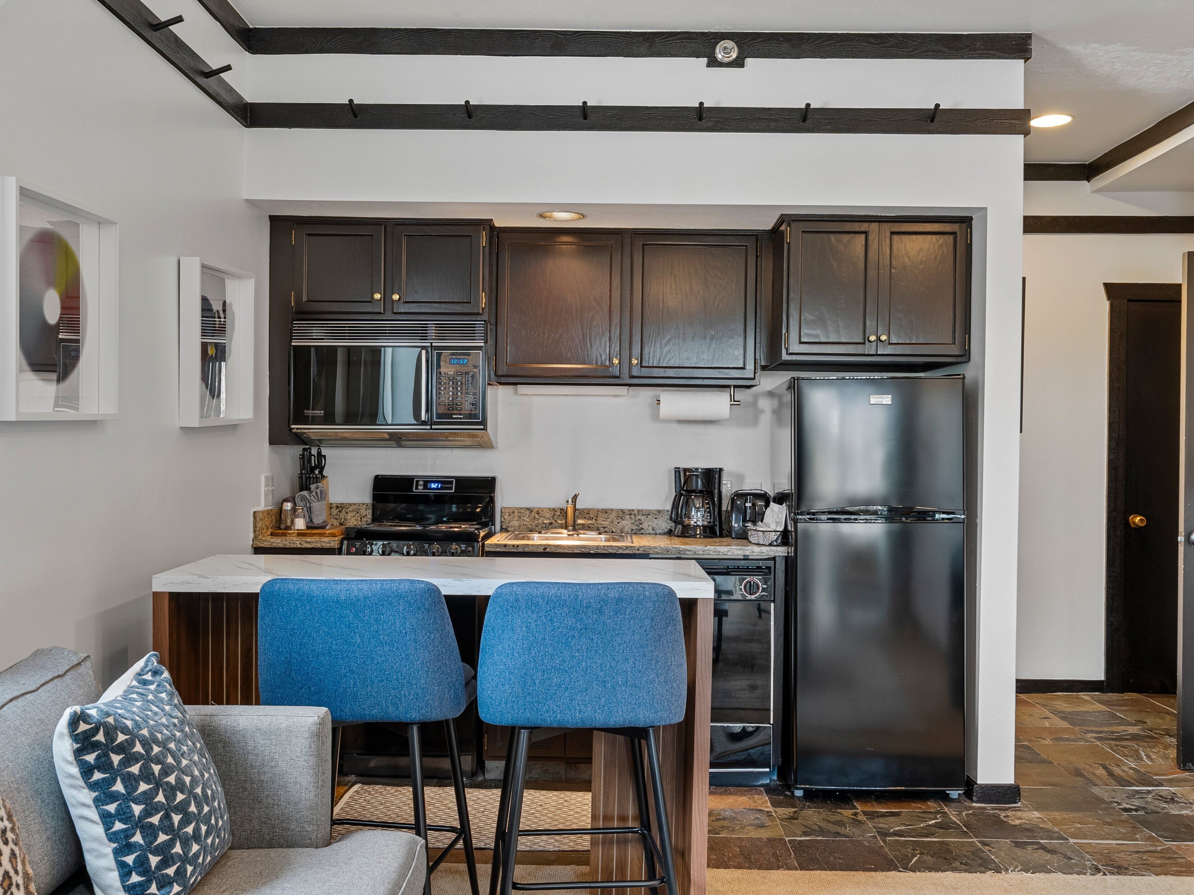 Une cuisine moderne avec des meubles de couleur sombre, un réfrigérateur en acier inoxydable, un four à micro-ondes et une cuisinière. On y trouve deux tabourets de bar bleus et un petit îlot central doté d’un évier.