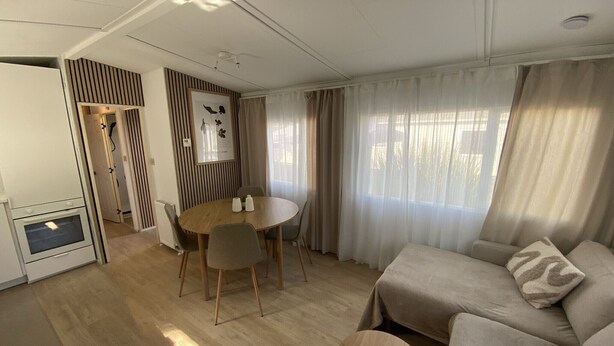 A modern living room with a sofa, a round wooden coffee table, a dining table with chairs, a kitchen area with an oven, and a large window with sheer curtains.