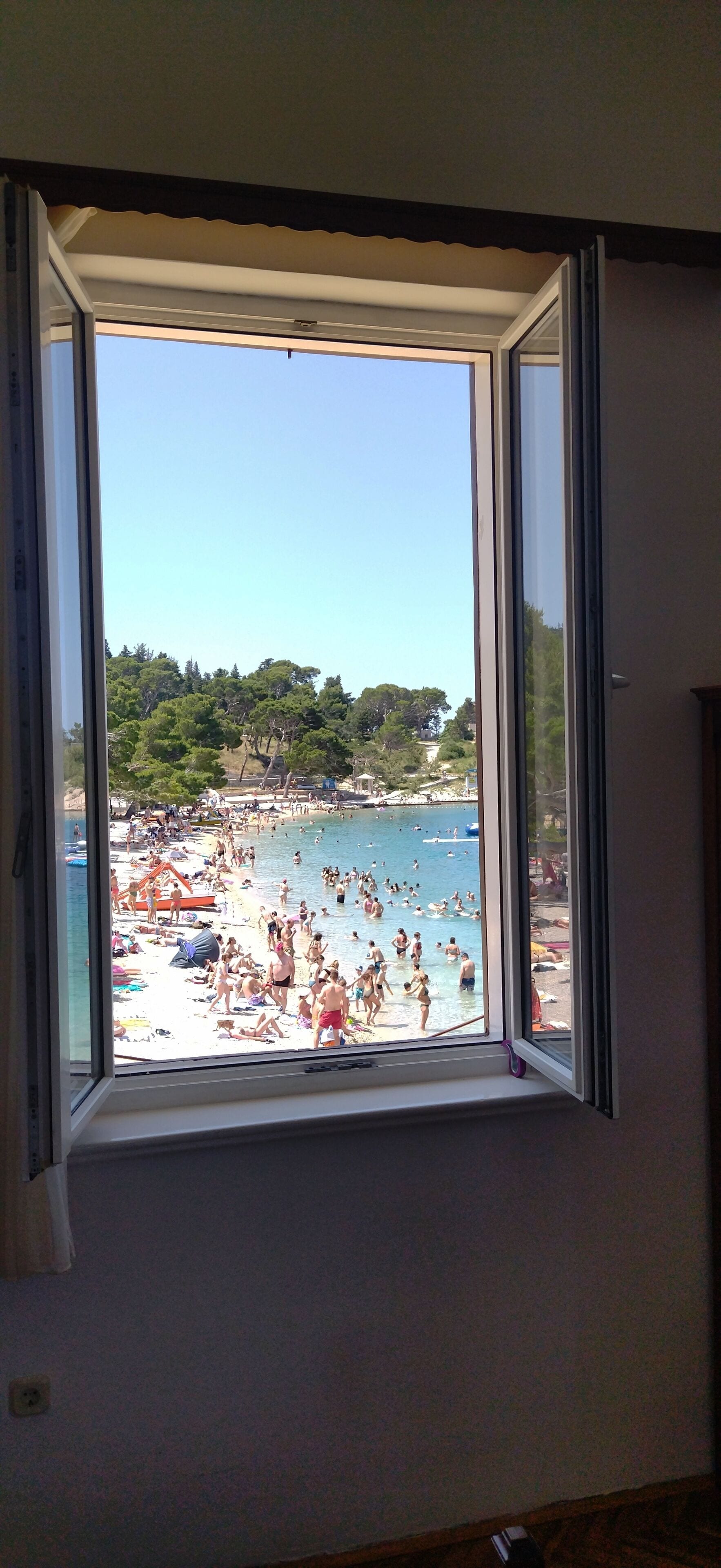 A room with a large window overlooking a crowded beach with people swimming and sunbathing.