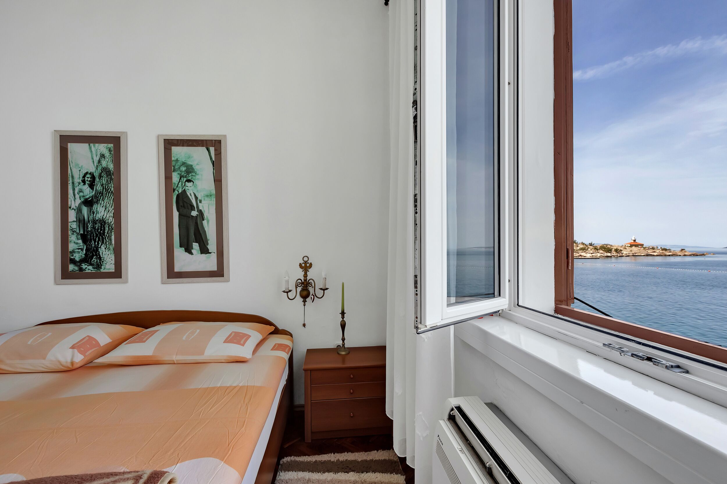 Une chambre à coucher comprenant un lit, une table de chevet, une lampe fixée au mur et une fenêtre donnant sur la mer.