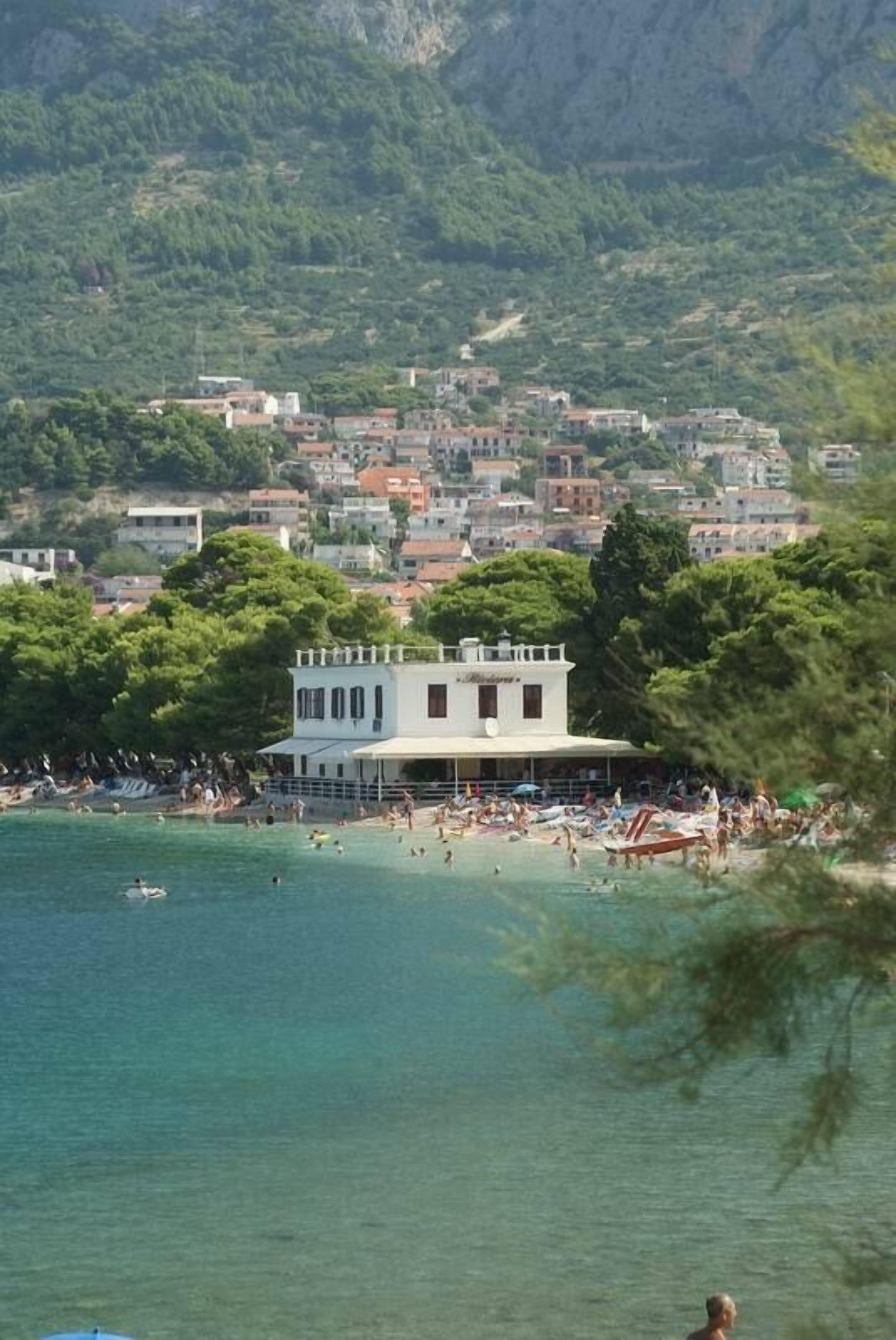 Un pueblo costero con una playa, un edificio blanco con balcón y un paisaje montañoso al fondo.