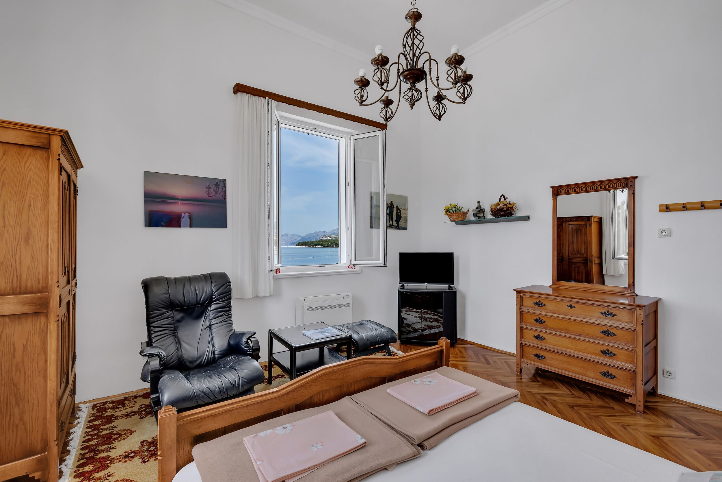 A bedroom with a bed, a wooden dresser, a black leather chair, a small table, a TV, and a window with a view.