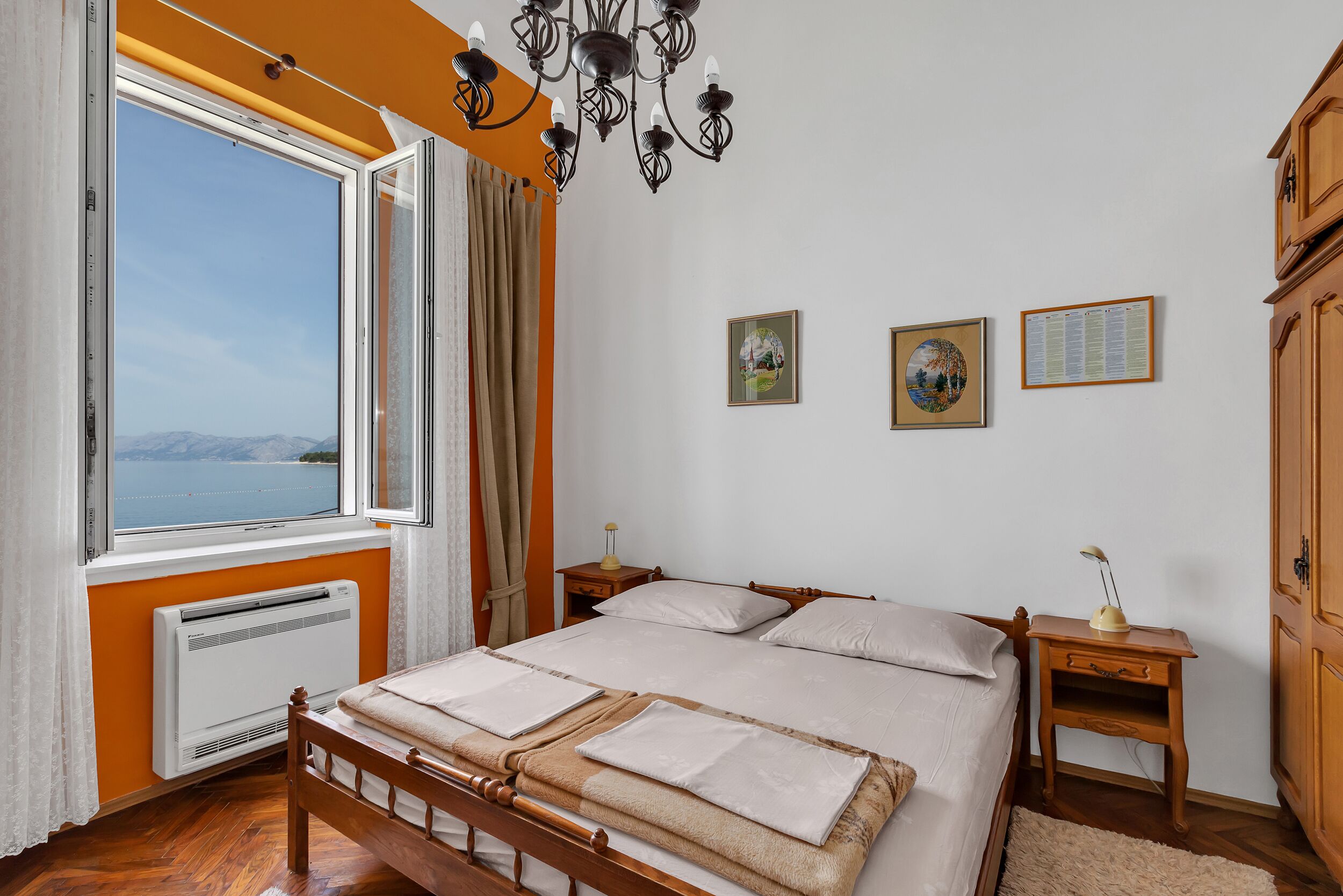 A bedroom with a bed, a nightstand, a heater, a window with curtains, and a view of the sea.