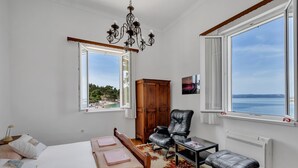 A bedroom with a bed, a chair, a table, a radiator, and large windows offering a view of the sea. - Beach rooms Riviera (Makarska)