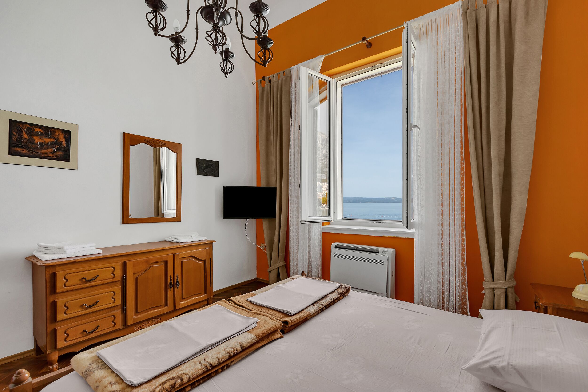 A hotel room with a bed, a wooden dresser, a flat-screen TV, a window with curtains, and a painting on the wall.
