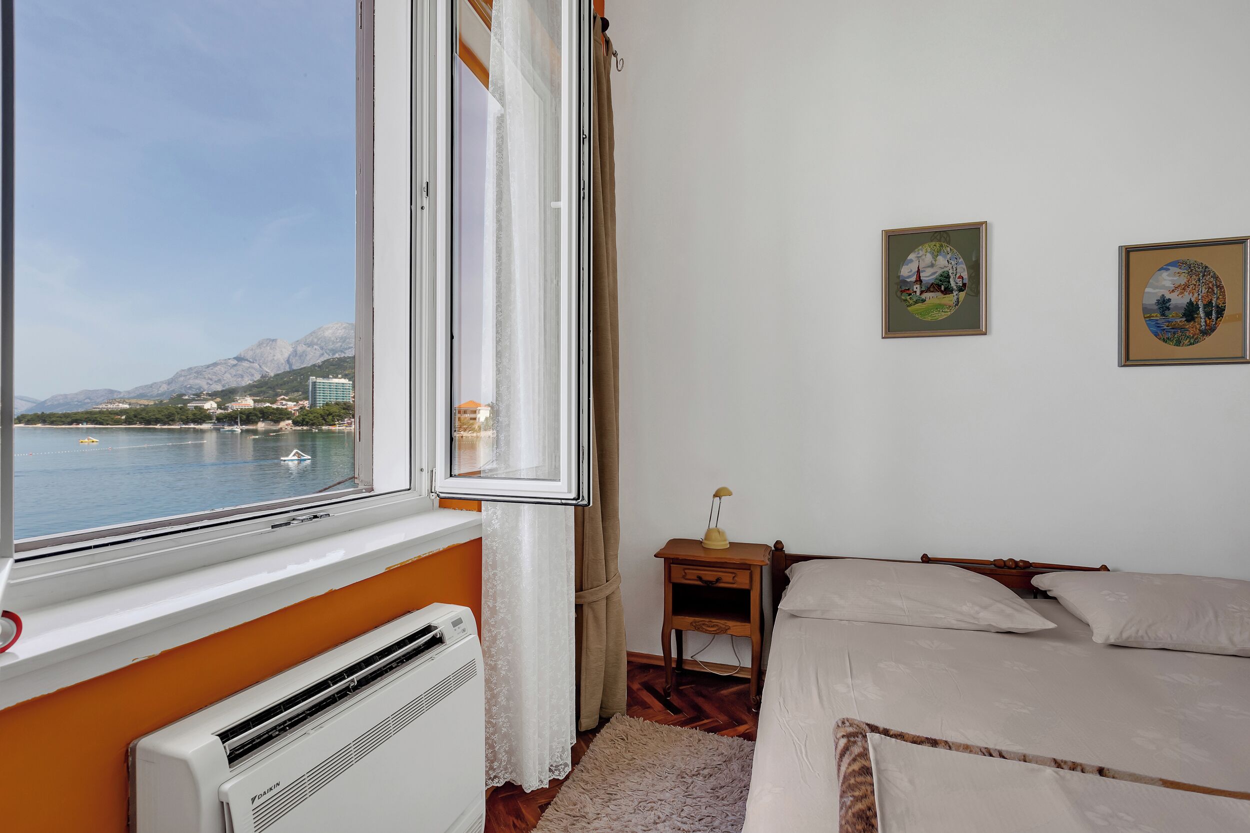 A room with a bed, a small bedside table, a heater, a window with a view of a body of water and mountains, and two framed pictures on the wall.