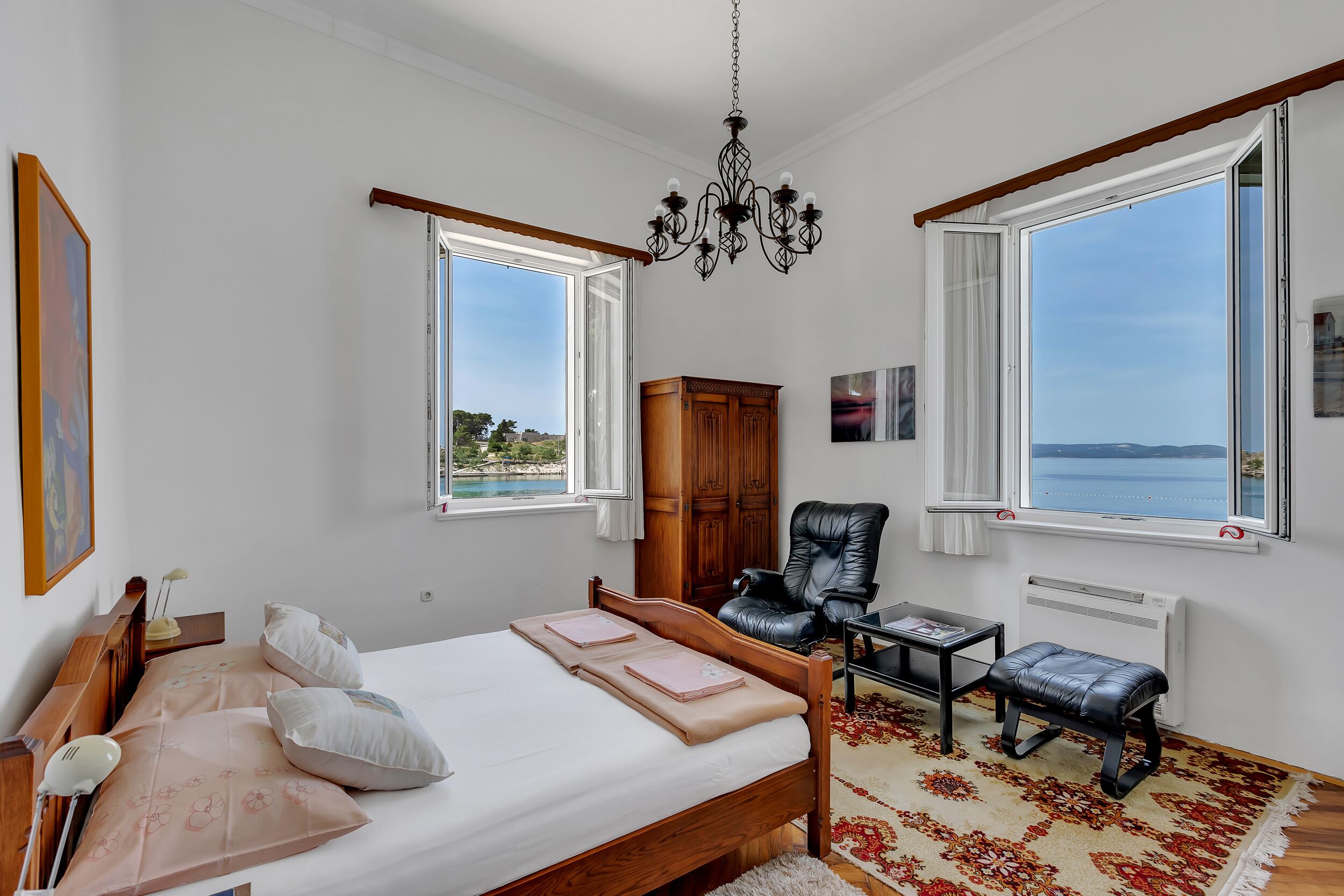 A bedroom with a bed, a chair, a desk, a heater, and large windows with a view.