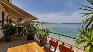 A balcony with a wooden table and chairs, potted plants, and a view of a calm sea and distant shoreline.