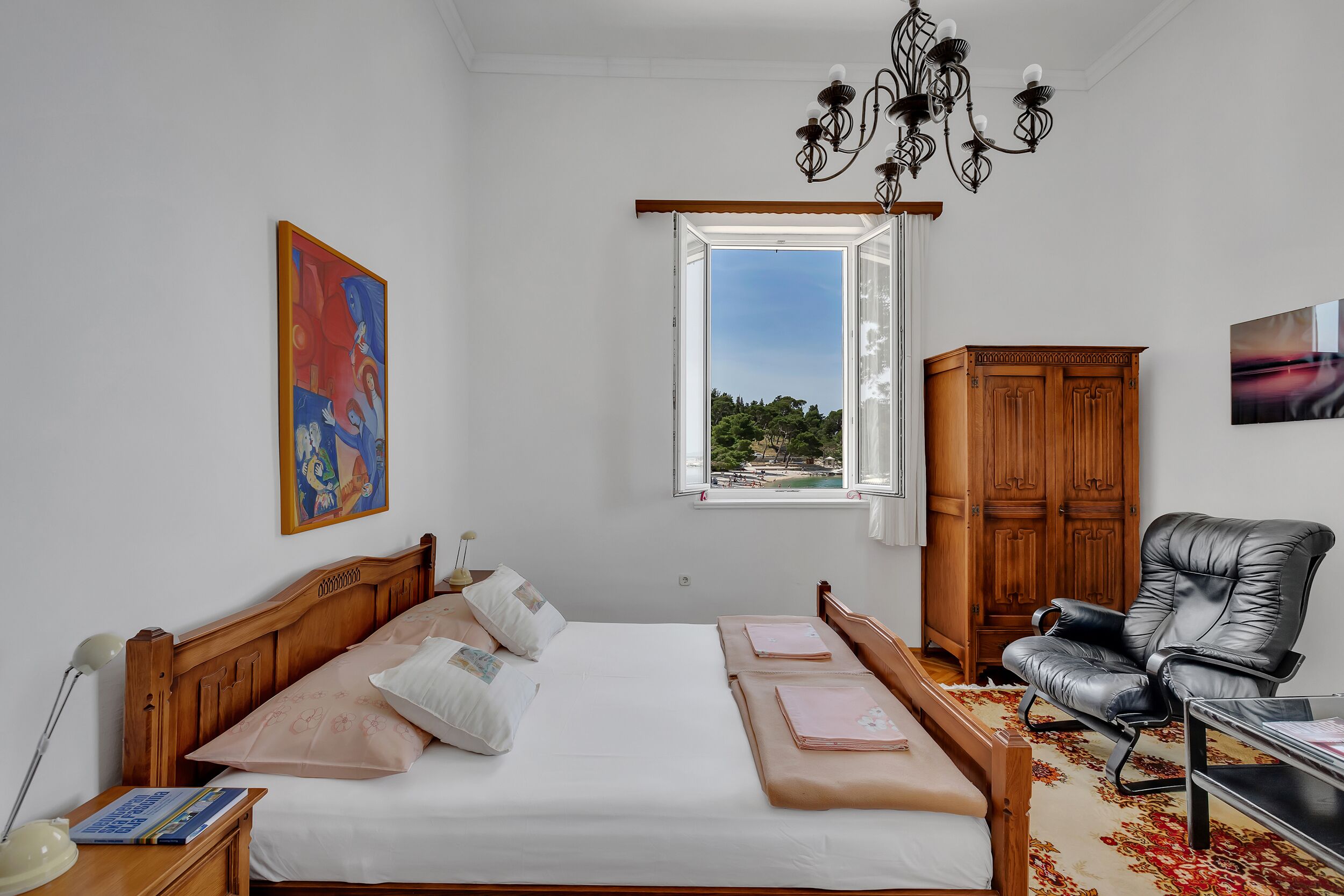 Une chambre à coucher avec un lit en bois, un fauteuil en cuir noir, une armoire en bois et une fenêtre donnant sur un paysage verdoyant.