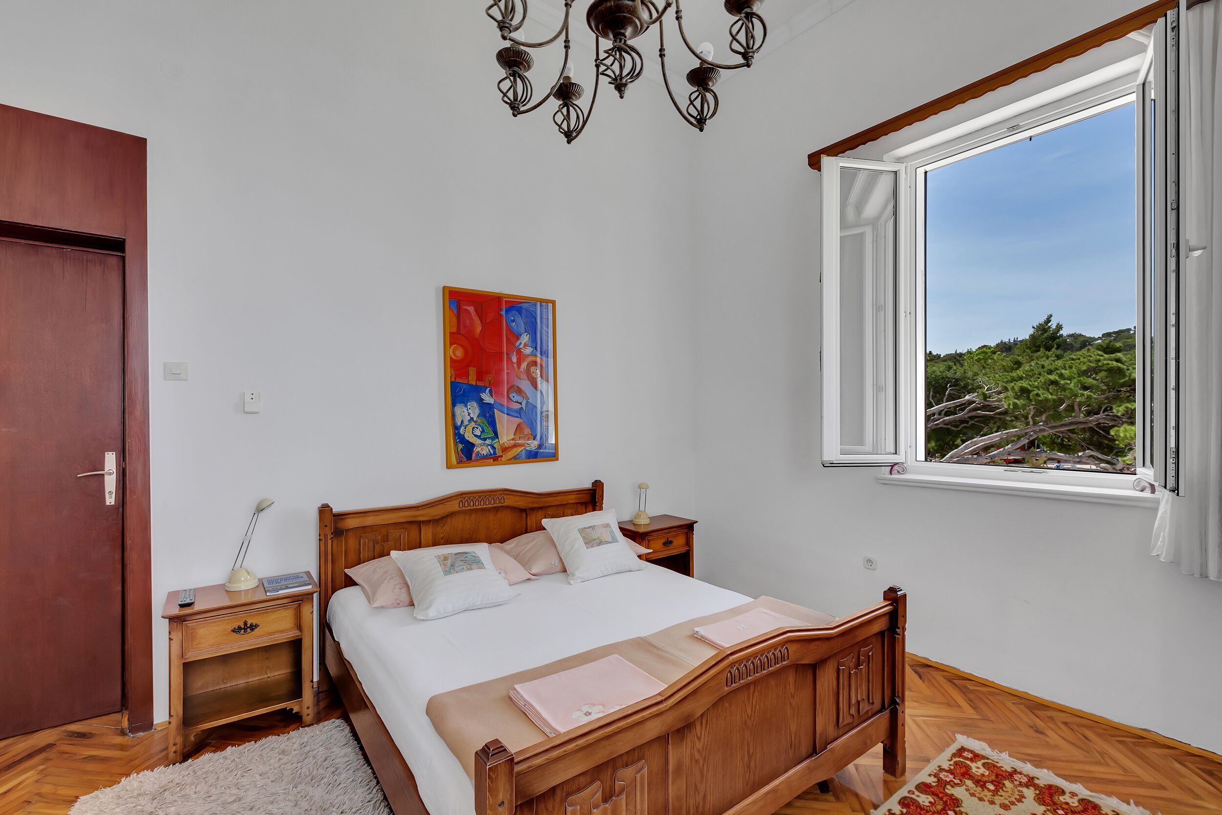 A bedroom with a wooden bed, a nightstand, a lamp, a painting, and a window with a view of greenery.