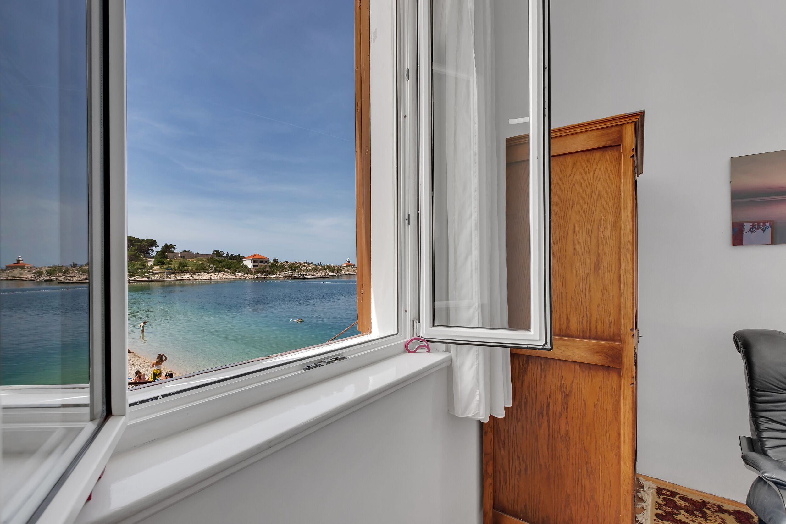 A room with a large window overlooking a beach and sea, a wooden door, and a chair.