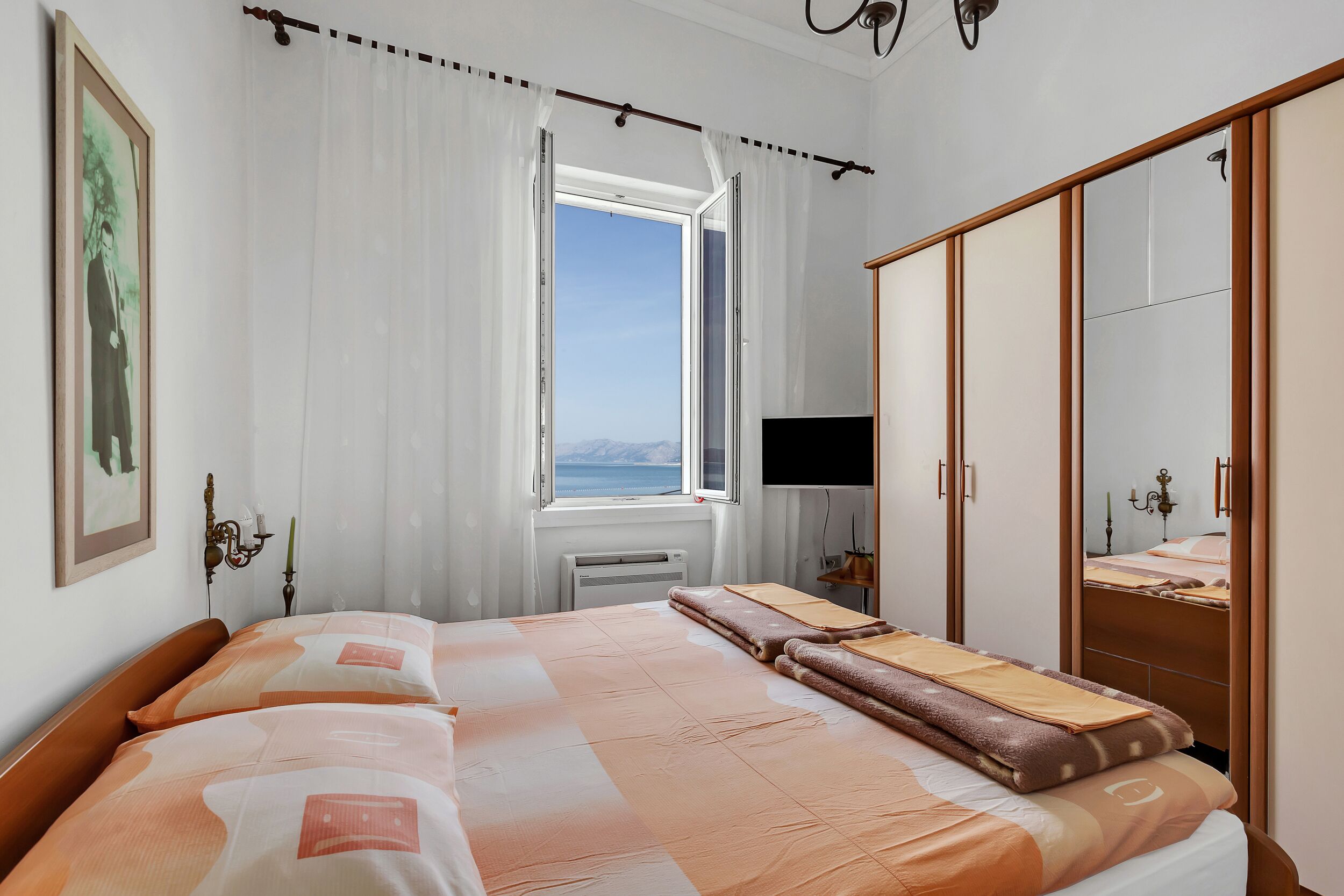 Ein Schlafzimmer mit Bett, Vorhängen am Fenster, Nachttisch, Spiegel und Kleiderschrank.