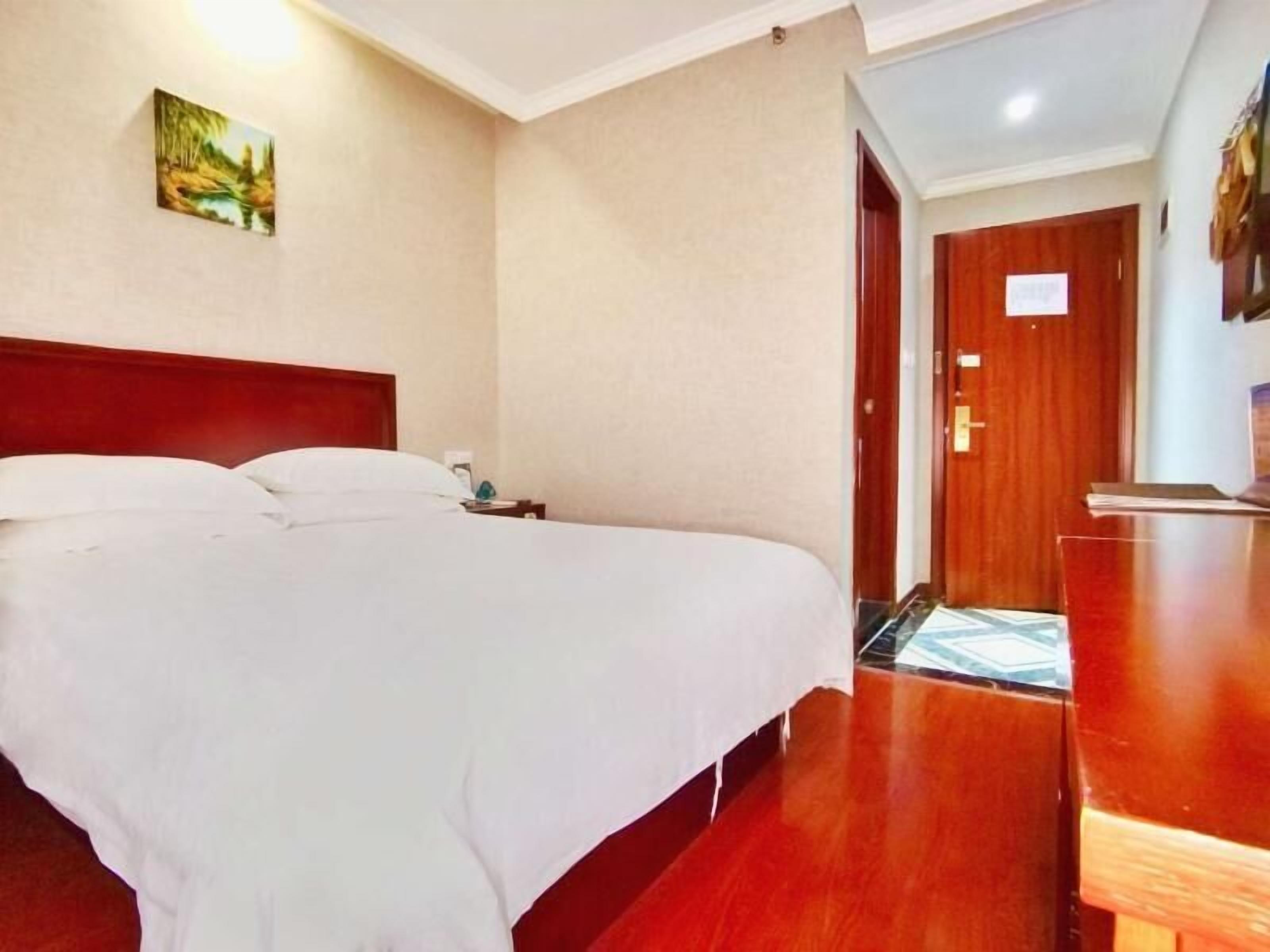 Una habitación de hotel con una cama grande, un aparador de madera, un cuadro en la pared y una puerta que da a otra habitación.