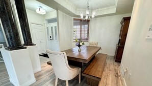 Een eetruimte met een houten tafel, beklede witte stoelen en een bank. Daarboven hangt een kroonluchter en er is een raam met jaloezieën.