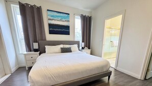 Een slaapkamer met een groot bed, een nachtkastje, een lamp, een schilderij aan de muur en uitzicht buiten het raam.