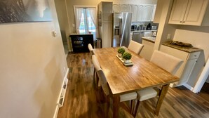 A dining area with a wooden table, chairs, and a small plant centerpiece. There is a kitchen in the background with white cabinets and appliances. - TC TWO OAKS Home / Minutes walk to Downtown & Beach (Traverse City)