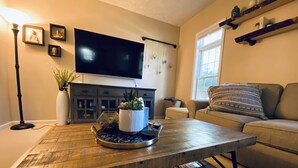 A living room with a wooden coffee table, a flat-screen TV mounted on the wall, a sofa, and a window. - TC TWO OAKS Home / Minutes walk to Downtown & Beach (Traverse City)