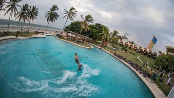 A person wakeboarding in a large pool with palm trees and a beach in the background.