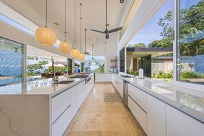 A modern kitchen with a central island, pendant lighting, and large windows offering views of the outdoors. - Ombak Luxury House- 5 bedroom Home / Concierge (Uvita)