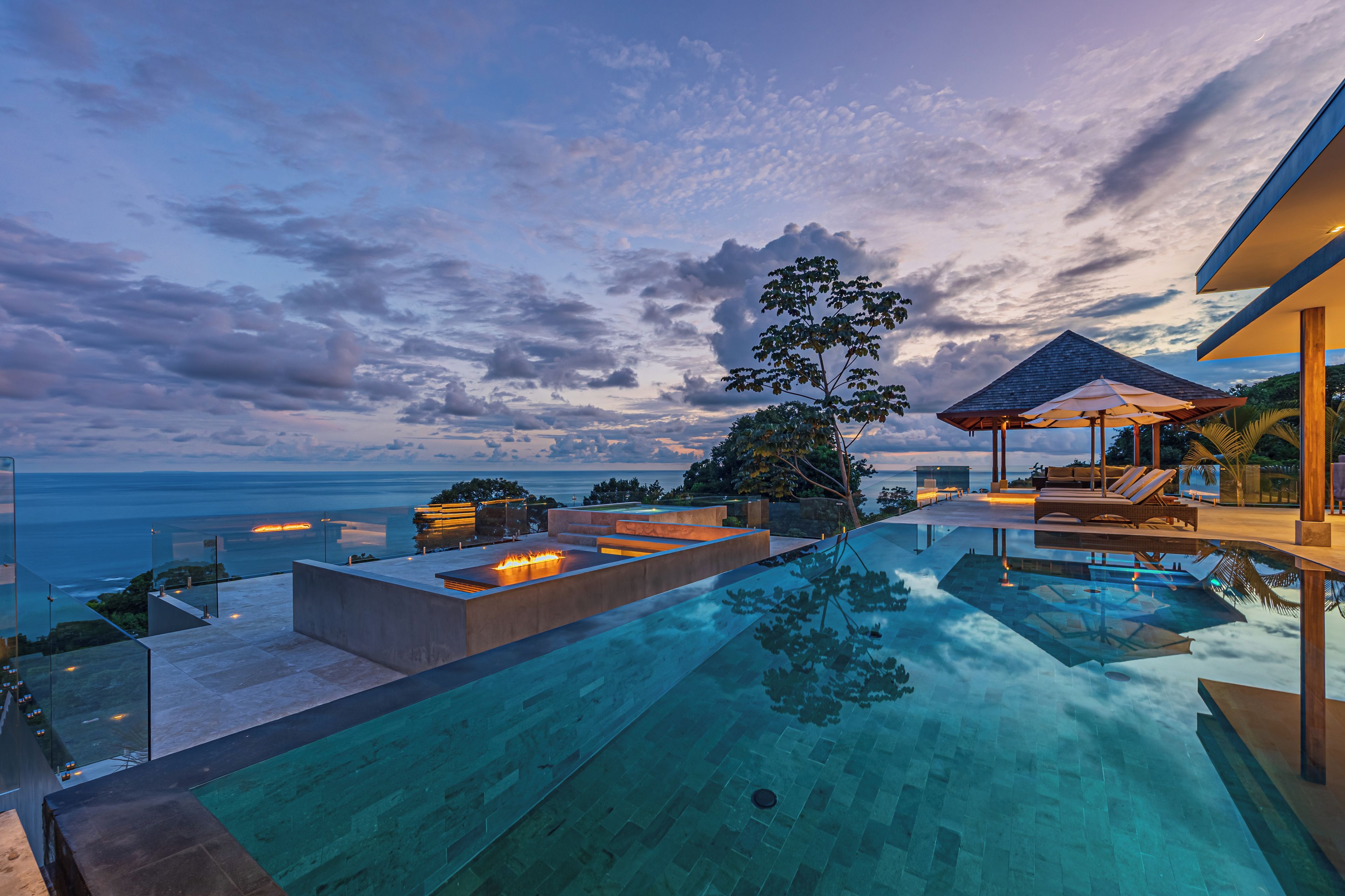 A modern infinity pool with a clear view of the ocean, a fire feature, and a lounge area with a pergola.