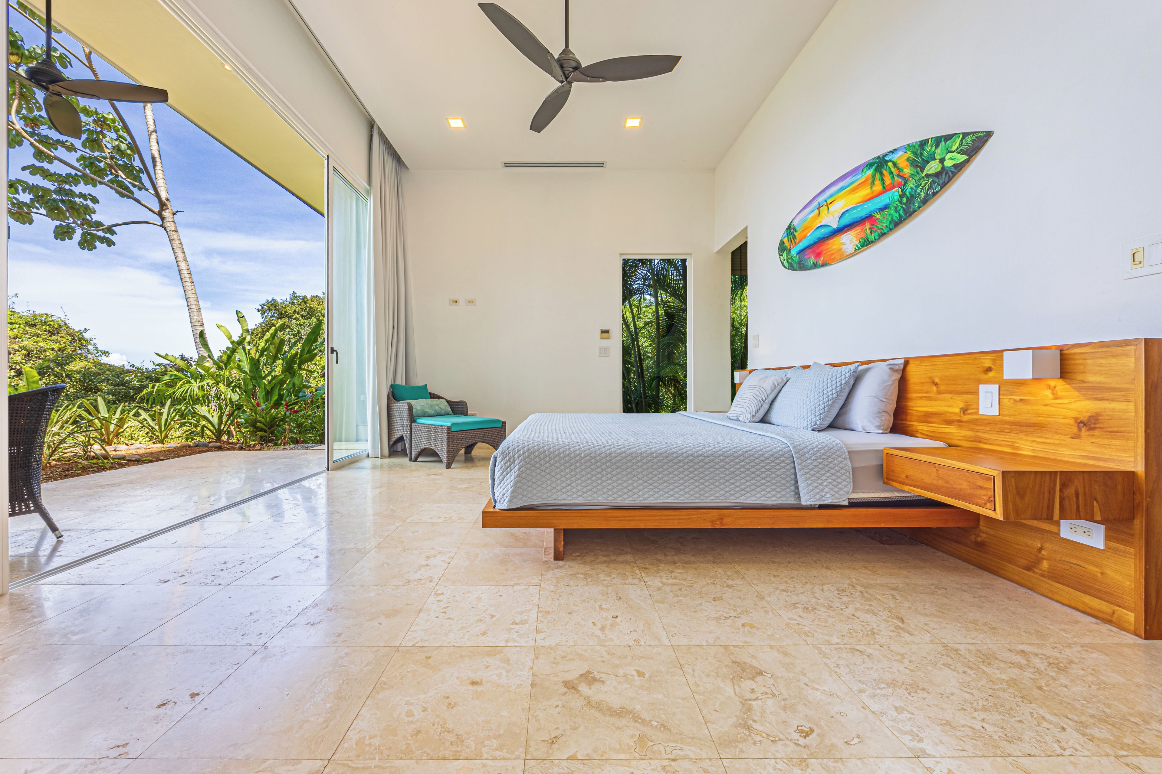 A modern bedroom with a large bed, a ceiling fan, and a view of greenery outside.