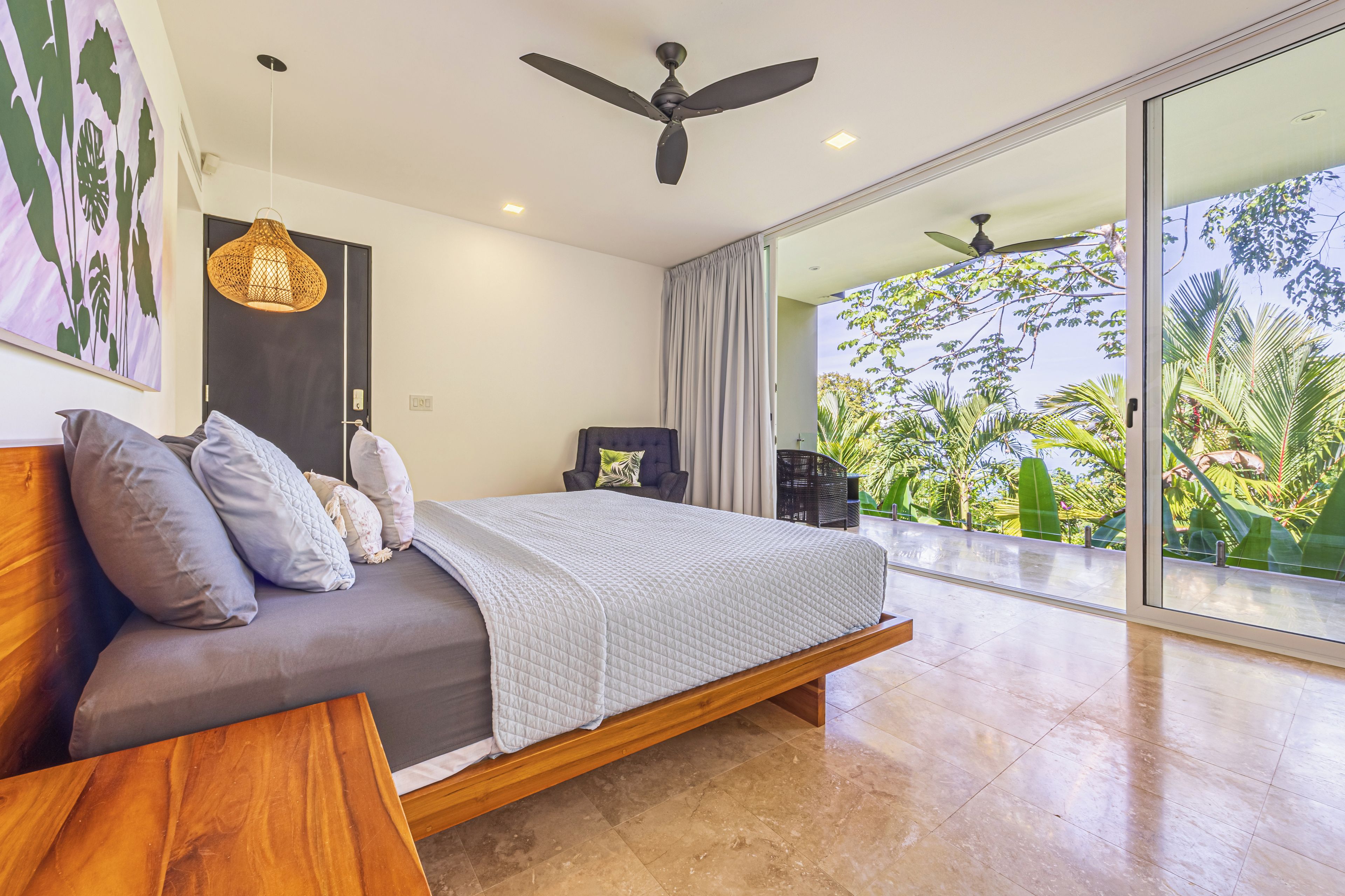 A modern bedroom with a bed, a wooden nightstand, a ceiling fan, and a view of lush greenery through a large sliding glass door.
