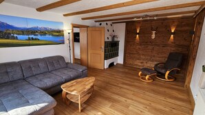 Un salon chaleureux avec un canapé gris, une table basse en bois et un fauteuil à bascule. On aperçoit une cheminée avec un manteau de couleur verte, et une grande fenêtre offrant une vue panoramique sur les montagnes et un lac.