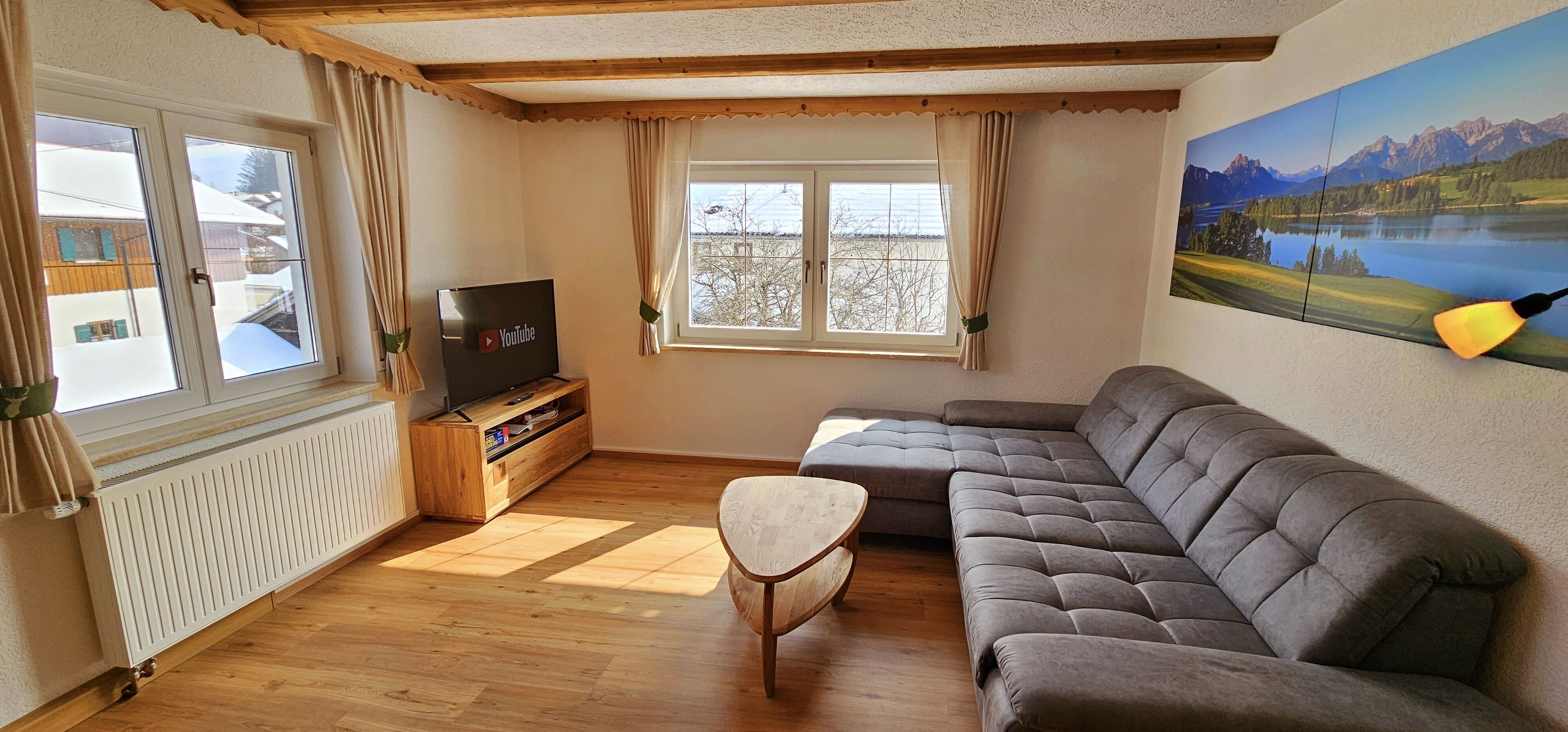 A cozy living room with a sectional sofa, a small wooden coffee table, a TV on a wooden stand, and a large window with curtains.