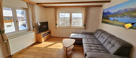 A cozy living room with a sectional sofa, a small wooden coffee table, a TV on a wooden stand, and a large window with curtains.