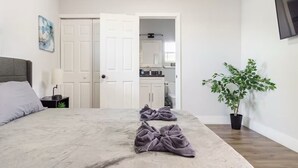 Un dormitorio con cama, mesita de noche, planta y baño visible a través de una puerta abierta.
