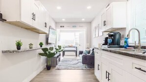 Una cocina estrecha con armarios blancos, una encimera y un fregadero. En el fondo, hay una zona de comedor con una mesa y sillas.