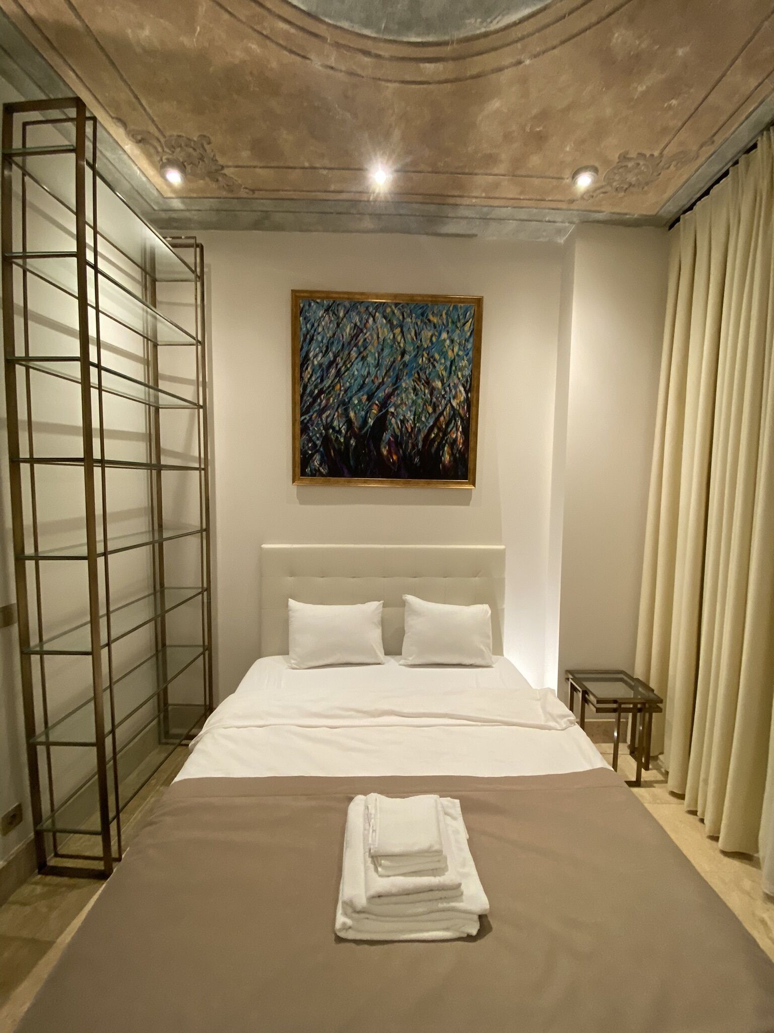 Una habitació d'hotel amb un llit gran, un quadre a la paret, una separació de vidre i una taula petita.