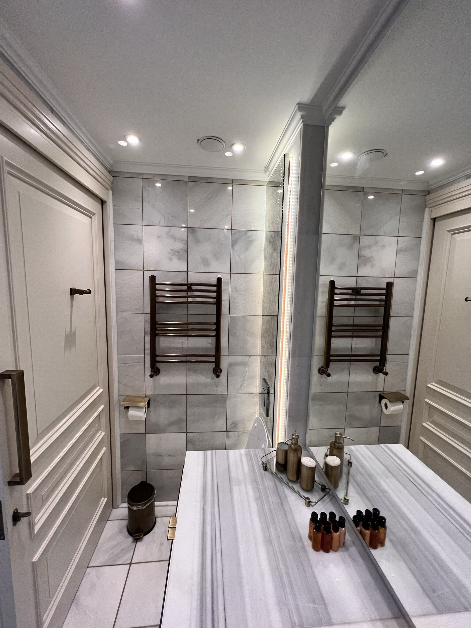 A bathroom with a large mirror, two towel racks, a sink with toiletries, and a trash can.