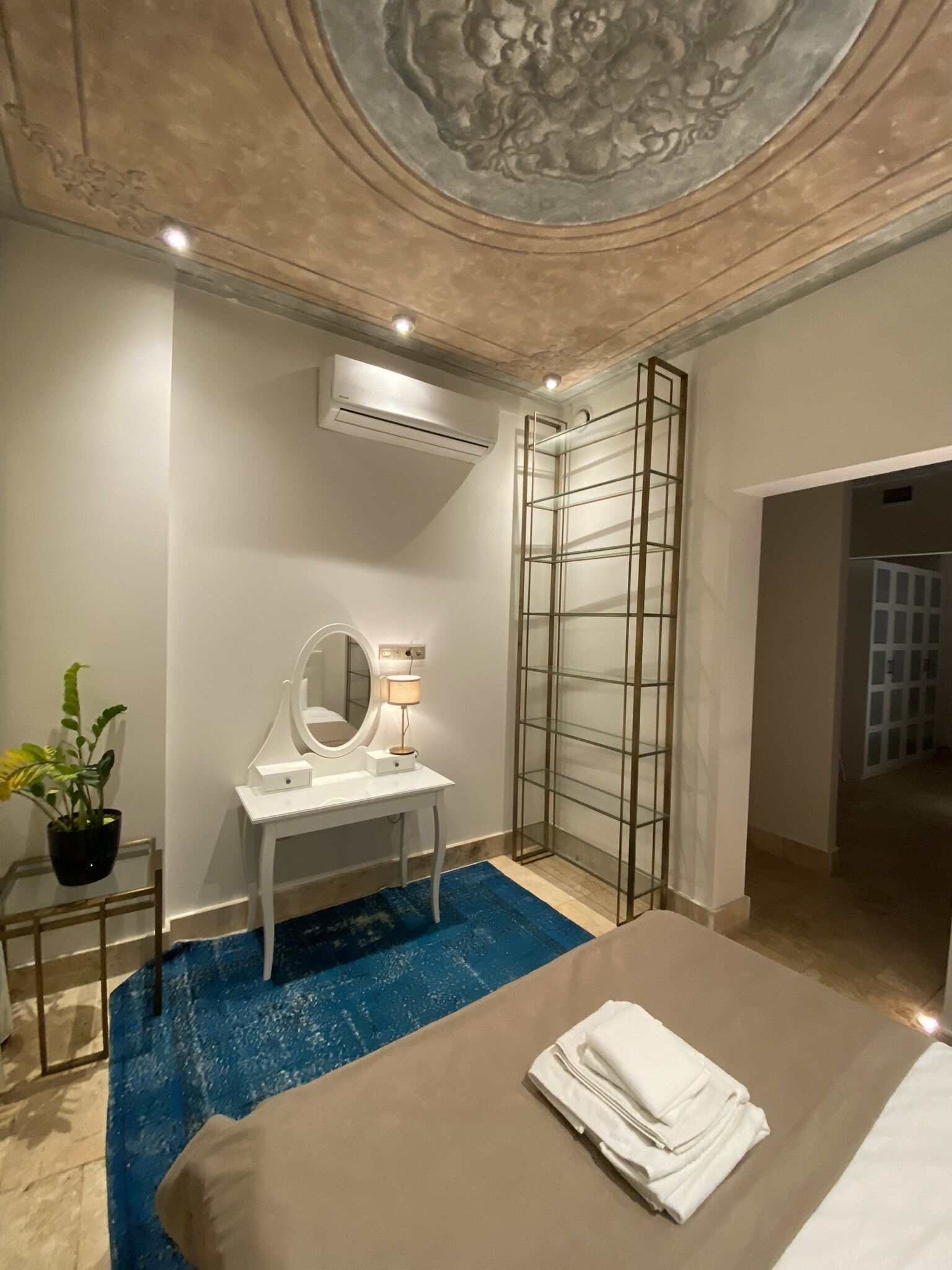 Une chambre d’hôtel moderne avec un lit, une coiffeuse avec un miroir, une plante en pot et un tapis bleu.