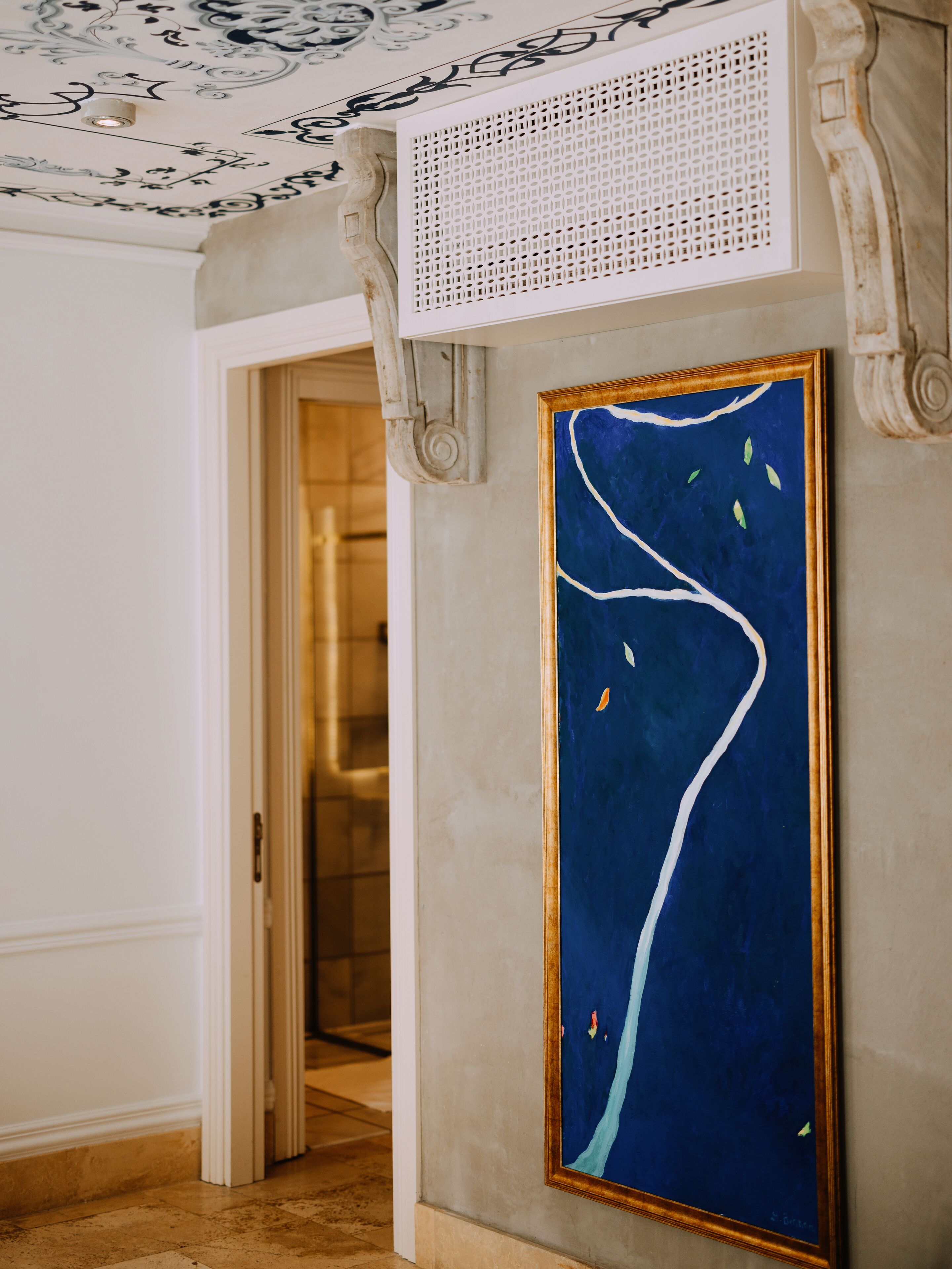 A hallway with a blue abstract painting, a patterned ceiling, and a doorway leading to another room.
