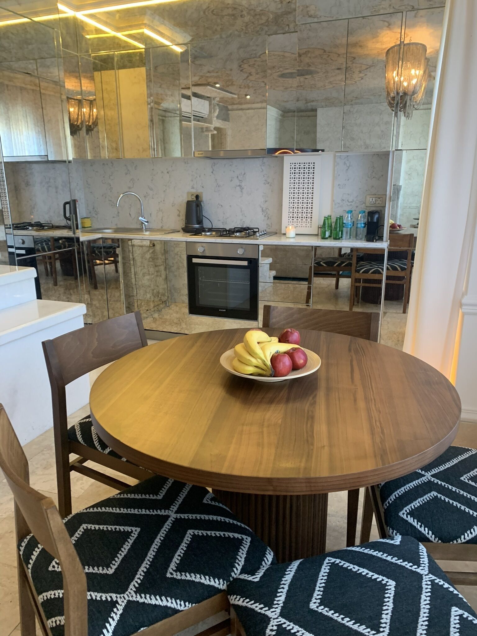A modern kitchen with a round wooden dining table, chairs with patterned cushions, and a fruit bowl.