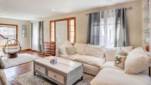 Una sala de estar con un sofá blanco, una mesa de centro y una silla. Se observa una puerta con ventana y cortinas.